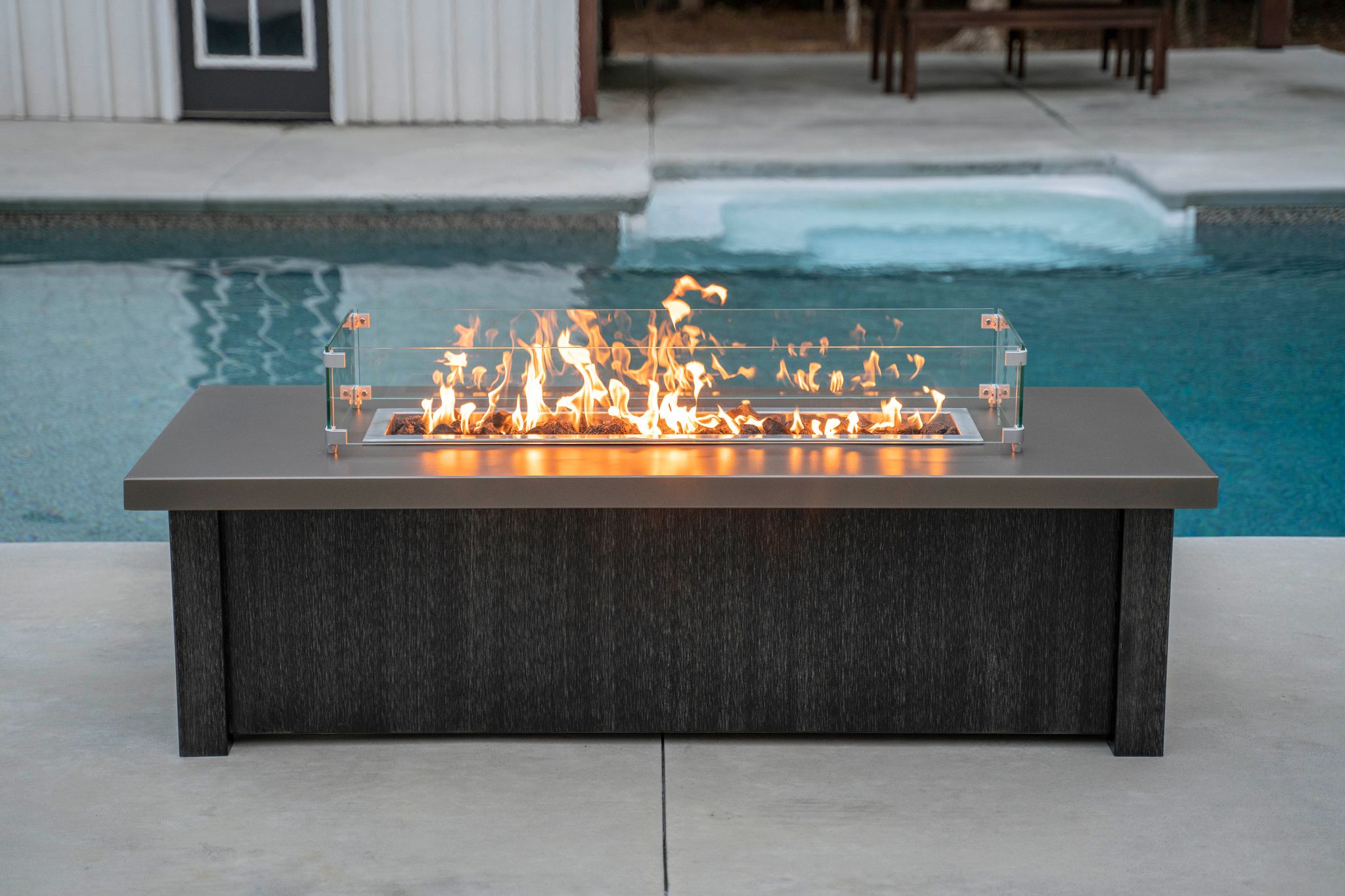 A fire pit table with wind shield sits by a pool.
