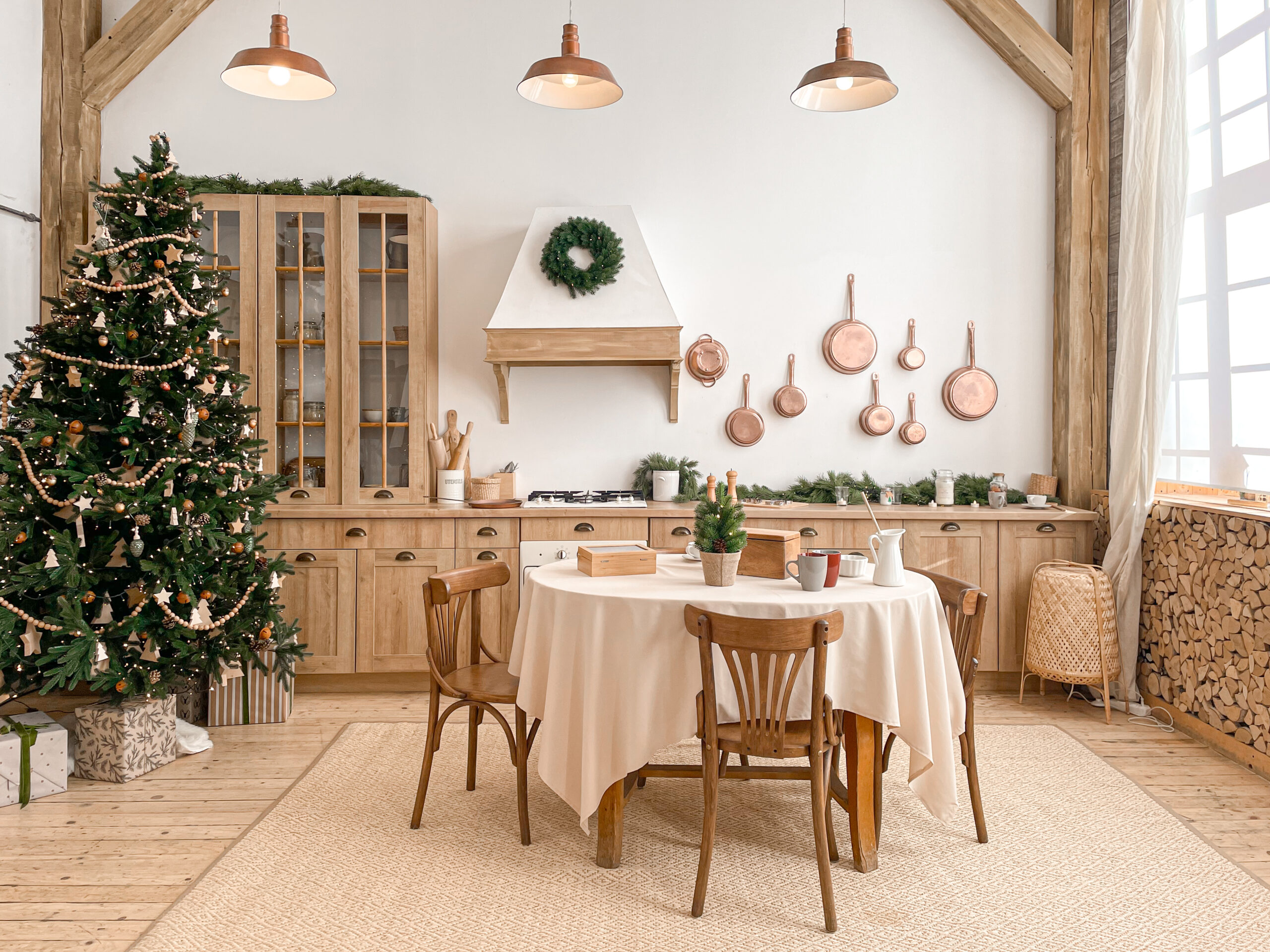 Modern, light, festive, cozy kitchen interior with Christmas and New Year decorations, kitchen table, utensils, copper pans on wall and big Christmas tree with presents, winter holidays concept